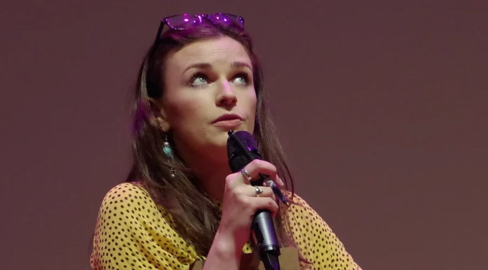 Aisling Bea Net Worth 2026: Career, Income and What We Know Aisling Bea performing stand-up comedy on stage holding a microphone and a clipboard