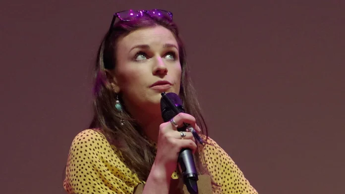 Aisling Bea performing stand-up comedy on stage holding a microphone and a clipboard