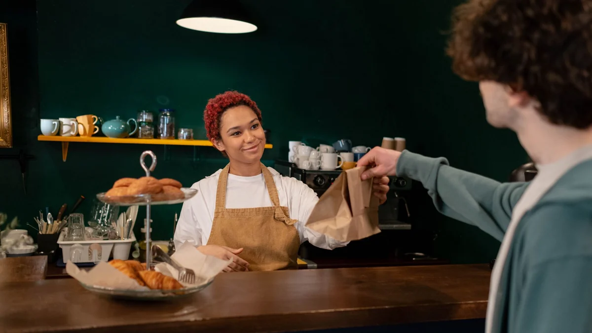 A cafe owner handing a paper bag of pastries to a customer across a wooden counter